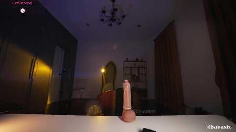 A dimly lit, modern bathroom with a beige countertop featuring a pink dildo. The room has a chandelier, a mirror, and a closed door. The atmosphere is intimate and slightly erotic.
