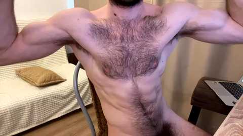 Video of a shirtless, muscular, hairy-chested man in a bedroom with a white bed, beige curtains, and a wooden chair.