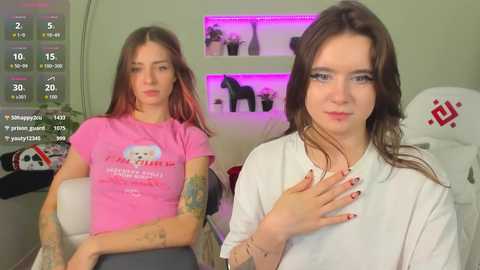 Video of a young woman with tattoos, wearing a pink shirt, sitting on a white chair. Another woman with long hair, wearing a white shirt, is standing beside her.