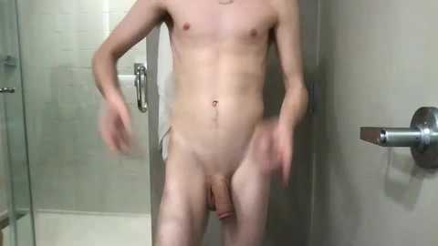 Video of a nude, light-skinned man with a small, flaccid penis standing in a modern bathroom with a glass shower door and silver fixtures.