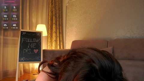 Video of a woman with long, dark hair lying on a beige couch, with a chalkboard sign reading \"Follow Me\" and a heart symbol beside her. Warm, dim lighting with a digital clock in the background.