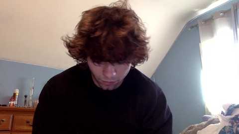 A video of a young man with curly brown hair, wearing a black t-shirt, bending over in a dimly lit, beige-walled room with a wooden dresser and a window with light streaming in.