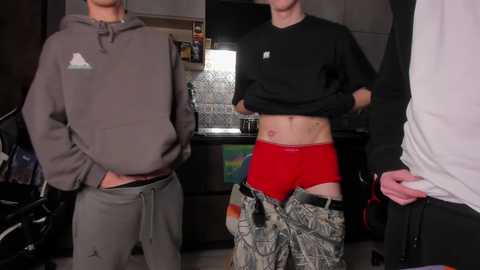 Video of three young men in casual attire. One wears a gray hoodie, another a black shirt, and the third has red shorts and camouflage pants. Background includes a flat-screen TV, shelves, and cluttered room.