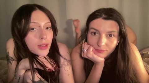 Video of two young women with pale skin, dark hair, and tattoos lying close together on a bed. They wear black tank tops, with one resting her chin on her hand.