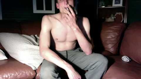 Video of a shirtless, bearded man smoking a cigarette, sitting on a brown leather couch with a blanket and pillows, in a dimly lit living room.