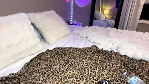 Video of a cozy bedroom with white fluffy pillows and blankets, a leopard-print blanket, and purple LED lights; a mirror reflects part of the room.