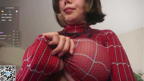 Video of a woman in a red Spider-Man costume, holding the sides of her top to reveal her bare chest. She has short, dark hair. Background shows a smartphone screen with a live stream.