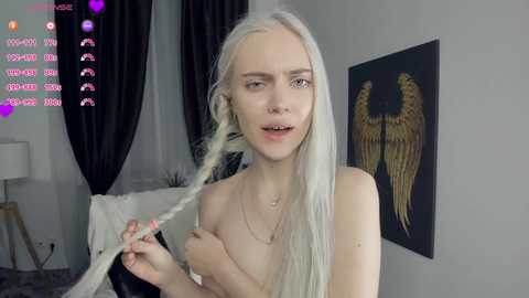 Video of a slender, pale-skinned, platinum blonde woman with long hair, topless, holding a strand of hair, in a modern, minimally decorated room with a dark, winged angel painting on the wall.