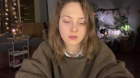 A video of a young, fair-skinned girl with long, messy blonde hair, wearing a brown sweater, sitting in a dimly lit bedroom with a bed, plants, and fairy lights in the background.