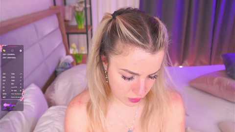 A video of a blonde woman with pigtails, wearing a strapless top, sitting on a bed with a white headboard, surrounded by pillows.