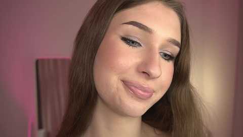 A video of a young woman with light skin, long brown hair, and a slight smile. She has well-defined eyebrows and wears black eyeliner. The background is a blurred, pinkish wall.