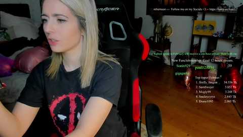 A video of a blonde woman in a black graphic tee, seated in a black gaming chair, streaming on Twitch. The room has a cozy, cluttered atmosphere with a wooden floor and gaming equipment.