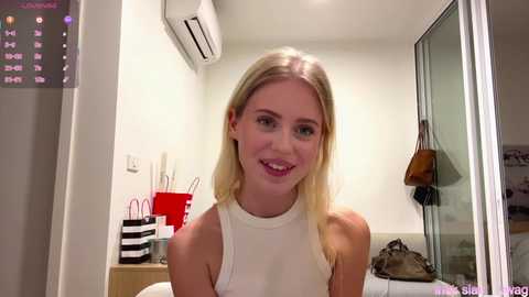 Video of a smiling blonde woman in a sleeveless white top, standing in a modern, minimalist room with white walls, an air conditioner, and a glass door.