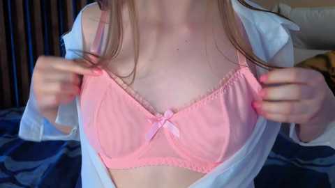 Video of a fair-skinned woman with long brown hair, wearing a pink lace bra with a bow and an open white shirt, standing against a dark wooden background.