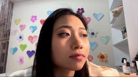 Video of an Asian woman with straight black hair, wearing a light-colored shirt, looking thoughtful in a room with colorful heart decorations on the wall.