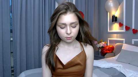 Video of a young woman with long brown hair, fair skin, and closed eyes, wearing a brown camisole, in a modern bedroom with gray curtains, white bed, and red heart decorations.