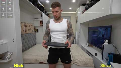 Video of a muscular, tattooed white man with short brown hair in a modern, minimalist bedroom. He wears a white tank top and black shorts, holding a black keyboard.