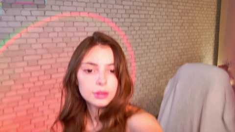 A video of a young woman with long brown hair, fair skin, and a neutral expression, sitting in front of a brick wall with a red light behind her.