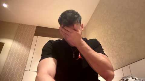 Video of a man with short dark hair, wearing a black t-shirt, covering his face with both hands, in a modern, beige-tiled bathroom with intricate patterned wallpaper.