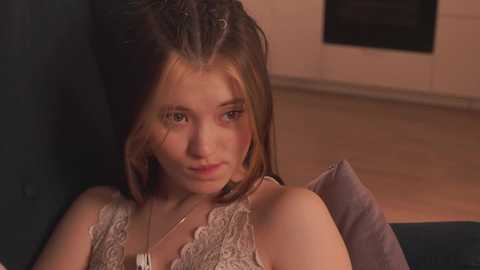 A video of a young woman with fair skin, light brown hair, and a delicate lace bra, reclining on a dark couch in a dimly lit room.