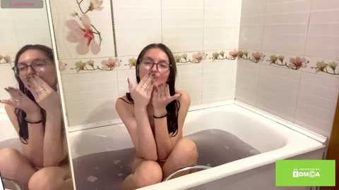 Video of a fair-skinned, slim woman with long dark hair, wearing glasses and a black top, sitting in a white bathtub, making a peace sign. The bathroom has floral-patterned tiles and a mirror reflection.
