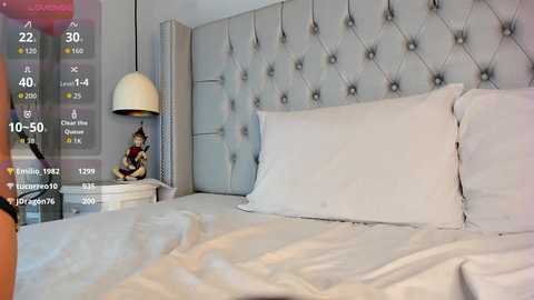 Video of a modern, minimalist bedroom with a tufted grey headboard, white bedding, a small white side table with a Buddha statue, and a white pendant lamp.