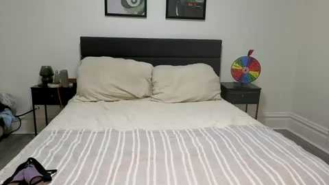 A video of a modern bedroom with a black headboard, beige pillows, and a white striped duvet on the bed. Nightstands with lamps flank the bed, and colorful artwork hangs on the wall.