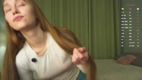 Video of a young, fair-skinned woman with long red hair, wearing a tight white top, standing in a dimly lit room with green curtains. She looks relaxed and slightly mischievous.