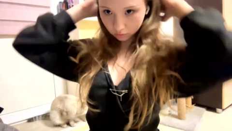 A video of a young woman with long, wavy brown hair, wearing a black V-neck top, adjusting her earphones in a casual indoor setting with a beige carpet and a beige wall.