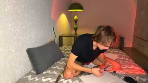 Video of a young woman with shoulder-length hair, kneeling on a bed with a gray and white chevron-patterned duvet, using a red marker on a sheet of paper. A green lamp with a black shade sits on a nightstand beside her.