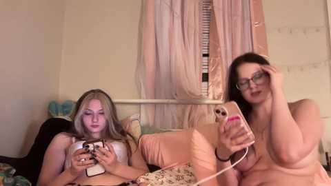 Video of two young women in a bedroom, one with long blonde hair and another with long dark hair and glasses, both topless, holding smartphones, in a cozy, cluttered room.