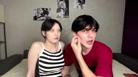 Video of two young people with pale skin and dark hair, wearing casual clothes. One adjusts the other's earpiece. Background: gray walls with black-and-white photos and a dark sofa.