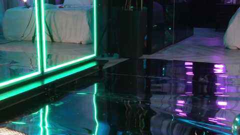 A video of a modern, dimly lit room with reflective, glossy black floor. Neon green and purple lights create a vibrant, futuristic ambiance. The background shows white-draped furniture, hinting at a formal event or party setting.