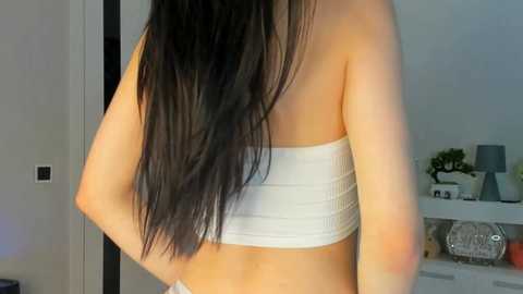Video of a woman's back, wearing a white bandeau top, long black hair, in a modern, minimalistic room with white walls and shelves.
