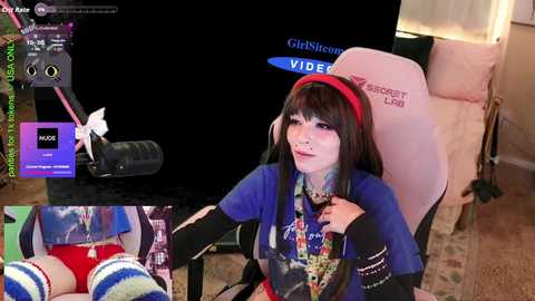 Video of a young Asian woman with pale skin, wearing a blue shirt, red headband, and headphones, sitting on a pink gaming chair. A video screen and a black microphone are visible in the background.