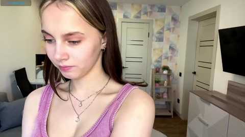 Video of a young woman with light skin and brown hair, wearing a pink tank top, standing in a colorful, pastel-themed room with floral wallpaper.