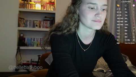 A video of a young woman with curly hair, wearing a black top, sitting on a bed in a cozy, dimly lit room. A video game screen displays her stats in the background.