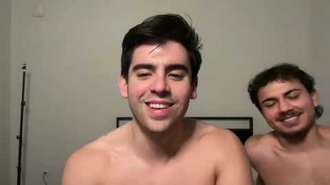 Media: Video of two shirtless young men with short dark hair, smiling at each other in a dimly lit room with a beige wall and a black framed picture.