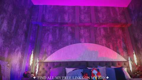 Video of a modern bedroom with a dark purple and pink lit ceiling, textured walls, a tufted gray headboard, and two modern lamps on either side of the bed, which is adorned with blue pillows.