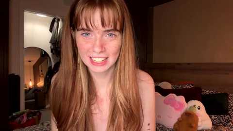 Video of a young, fair-skinned, red-haired girl with bangs smiling, surrounded by stuffed animals in a dimly lit bedroom.