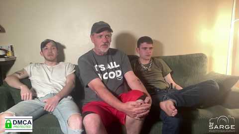 Video of three young men sitting on a green couch in a dimly lit living room. One wears a \"IS ALL GOD\" t-shirt, another a red shirt, and the third a black shirt.