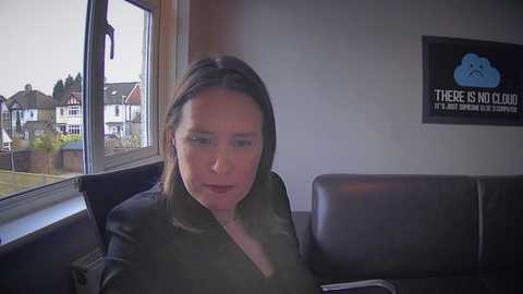 Video of a woman with light skin and shoulder-length brown hair, wearing a black jacket, sitting indoors by a window. She gazes outside at suburban houses.
