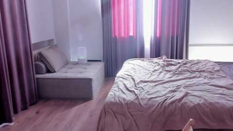 Video of a minimalist, modern bedroom with a queen-sized bed, beige bedding, purple curtains, and a white nightstand with a lamp and books.