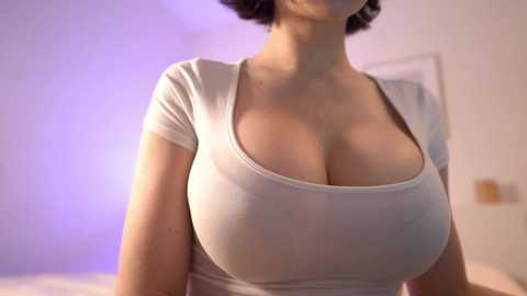 Video of a light-skinned woman with short dark hair, wearing a tight, white, scoop-neck shirt that accentuates her large breasts. Background features a blurred, softly lit room with purple lighting.