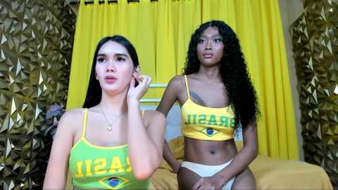 Video of two young women with light brown skin, wearing matching yellow Brazilian soccer jerseys and white panties, standing in a room with gold leaf walls and yellow curtains.