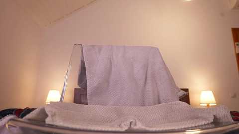 Video of a neatly made bed with a white, patterned duvet cover and two bedside lamps emitting warm light, set against a plain white wall.