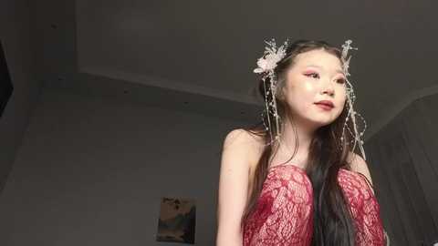 Video of an East Asian woman with long, straight black hair adorned with floral accessories. She wears a red, lace dress, standing indoors with a minimalist, white-walled background.