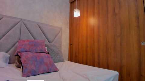 Video of a modern bedroom with a gray, tufted headboard, white bed linens, and a pair of pink and purple patterned pillows. A wooden closet door and a pendant light with a translucent shade are visible in the background.