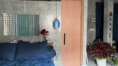 Video of a modern, cozy bedroom with a blue bed, wooden closet, gray concrete wall, and a potted red rose bouquet.