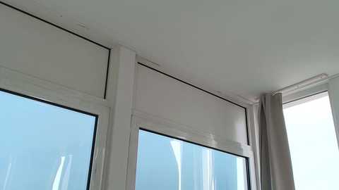 Video of a modern interior featuring large windows with white frames and sheer white blinds partially drawn. The room has a minimalist design with white walls and a neutral color palette.